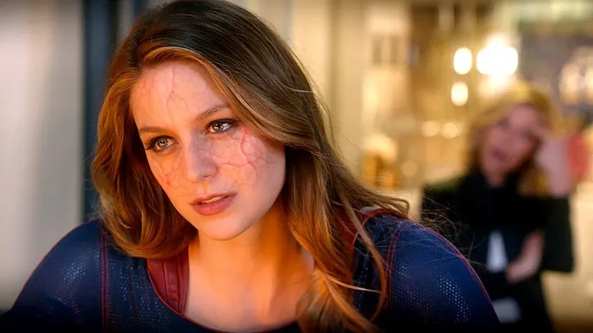 Kara con el traje de Supergirl y grietas rojizas en la cara, con una mujer rubia desenfocada al fondo